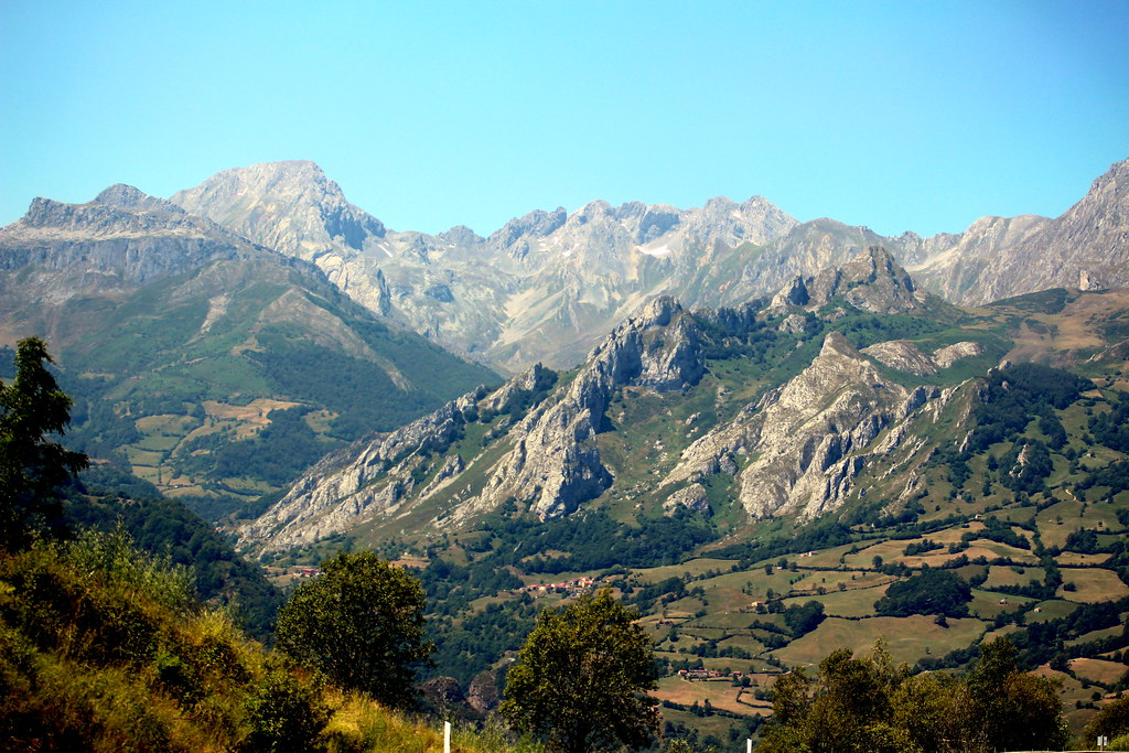 A wide shot of a mountainous landscape with rugged peaks, green valleys, and scattered houses, under a clear blue sky.