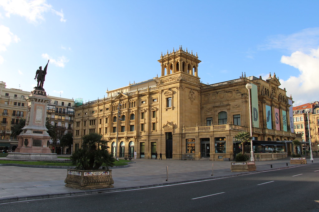 An architectural building with a statue in the square in front, located in a city with modern and historical buildings, under a bright sky.