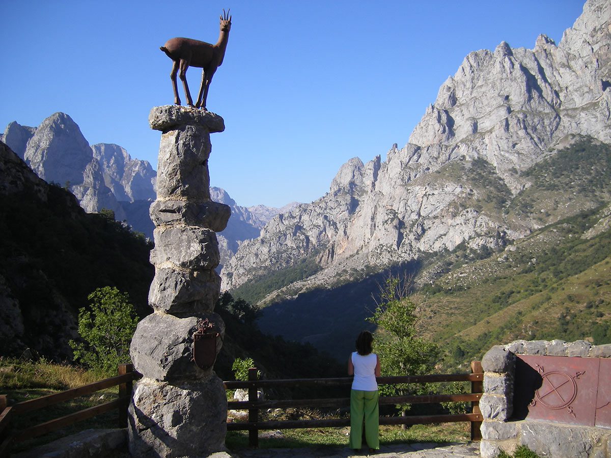 A person standing on a lookout with a view of rugged mountains, gazing at a rock sculpture of a goat placed atop a stone column.