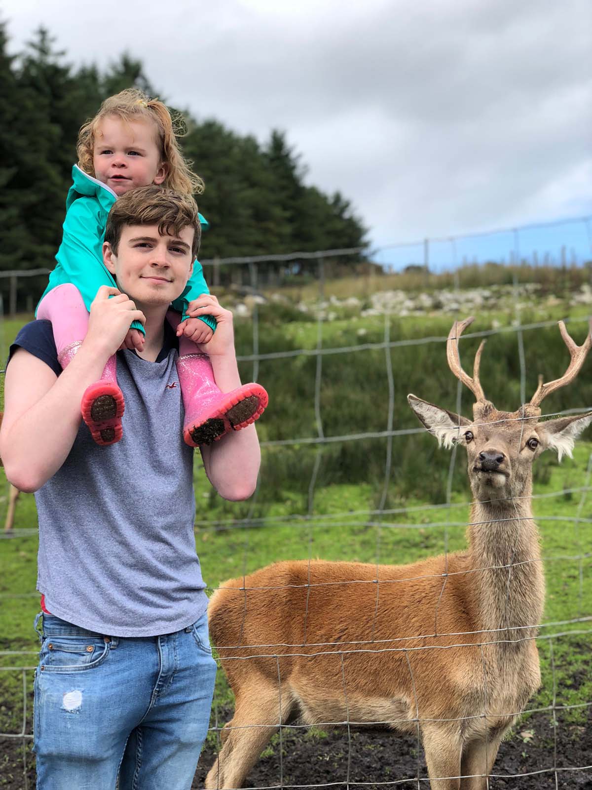 Young boy carrying a little girl on his shoulders, with a deer standing next to them in a fenced area, set against a lush green background.