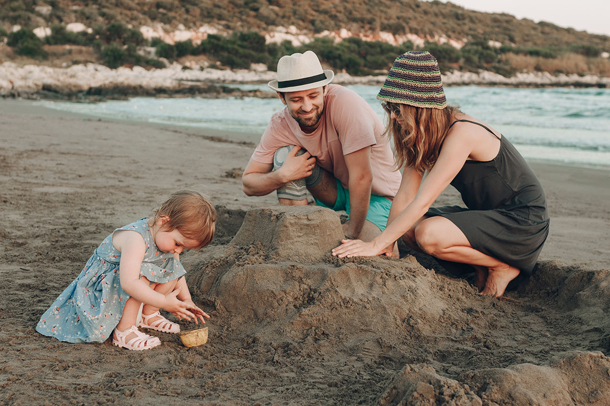 A family playing on the beach: a little girl wearing a blue dress and sandals is building a sandcastle, while her parents, dressed casually, kneel beside her and assist in the fun activity.