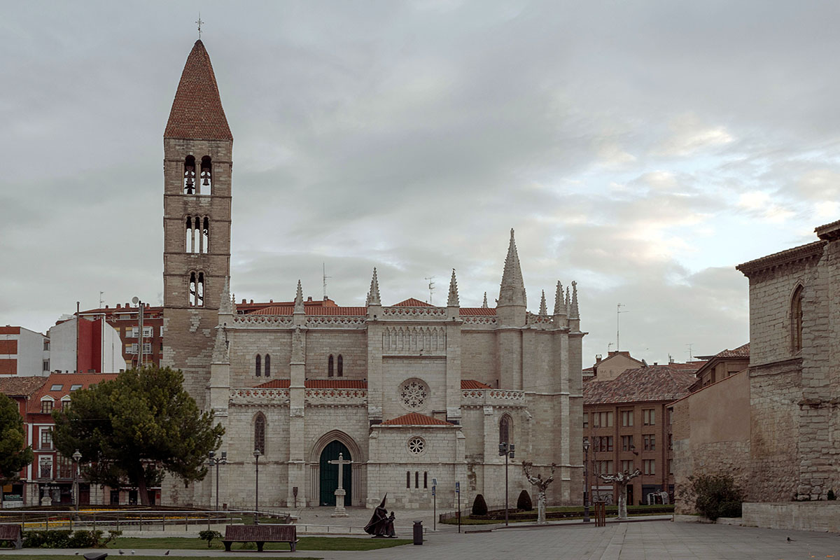 View of a historic church with a tall bell tower, spires, and a stone facade, located in a square surrounded by modern buildings.