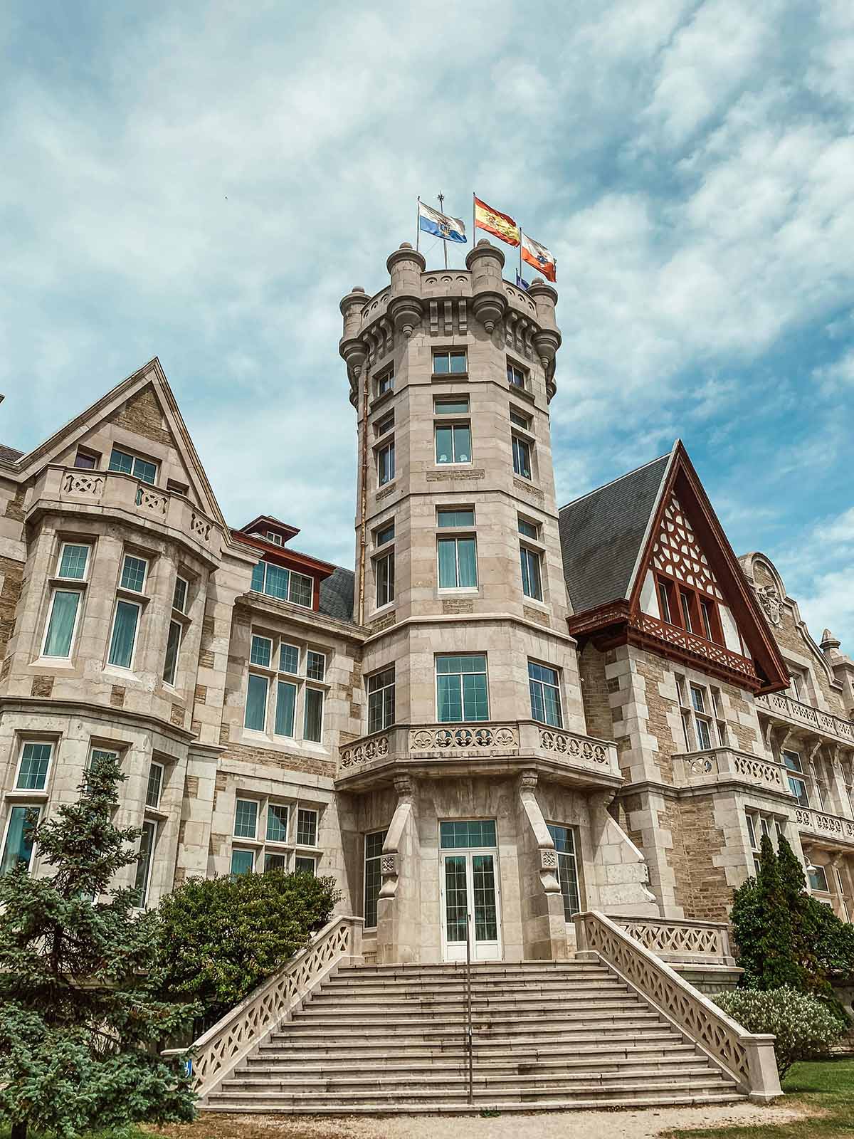 View of a grand building with a tower and flags at the top, featuring ornate stone architecture and a set of stairs leading to the entrance.