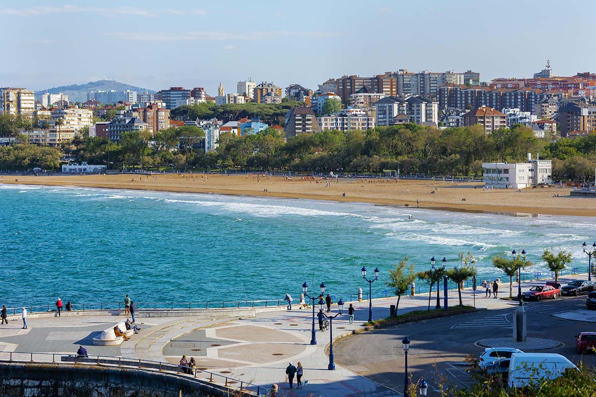View of a beach with golden sand and turquoise water, lined by a promenade with trees and benches, and residential buildings in the background under a bright sky.