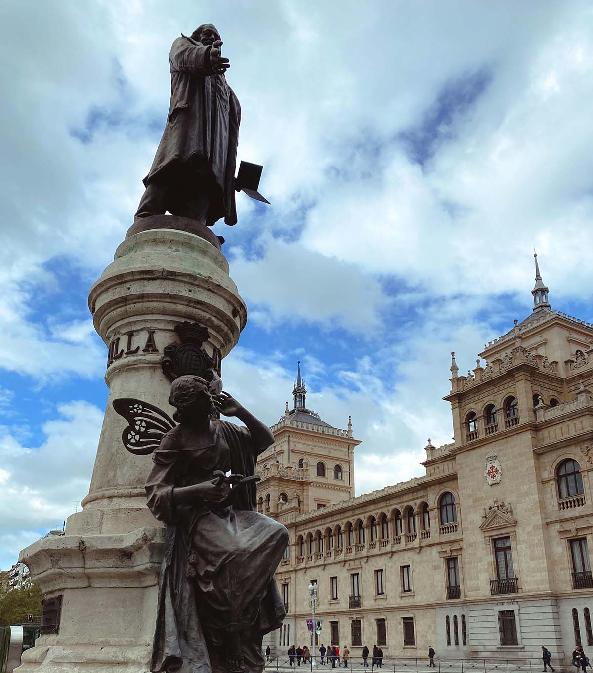 Statue of a prominent figure standing atop a pedestal, with a seated figure holding a butterfly at the base. The background shows an elegant historical building under a partly cloudy sky.