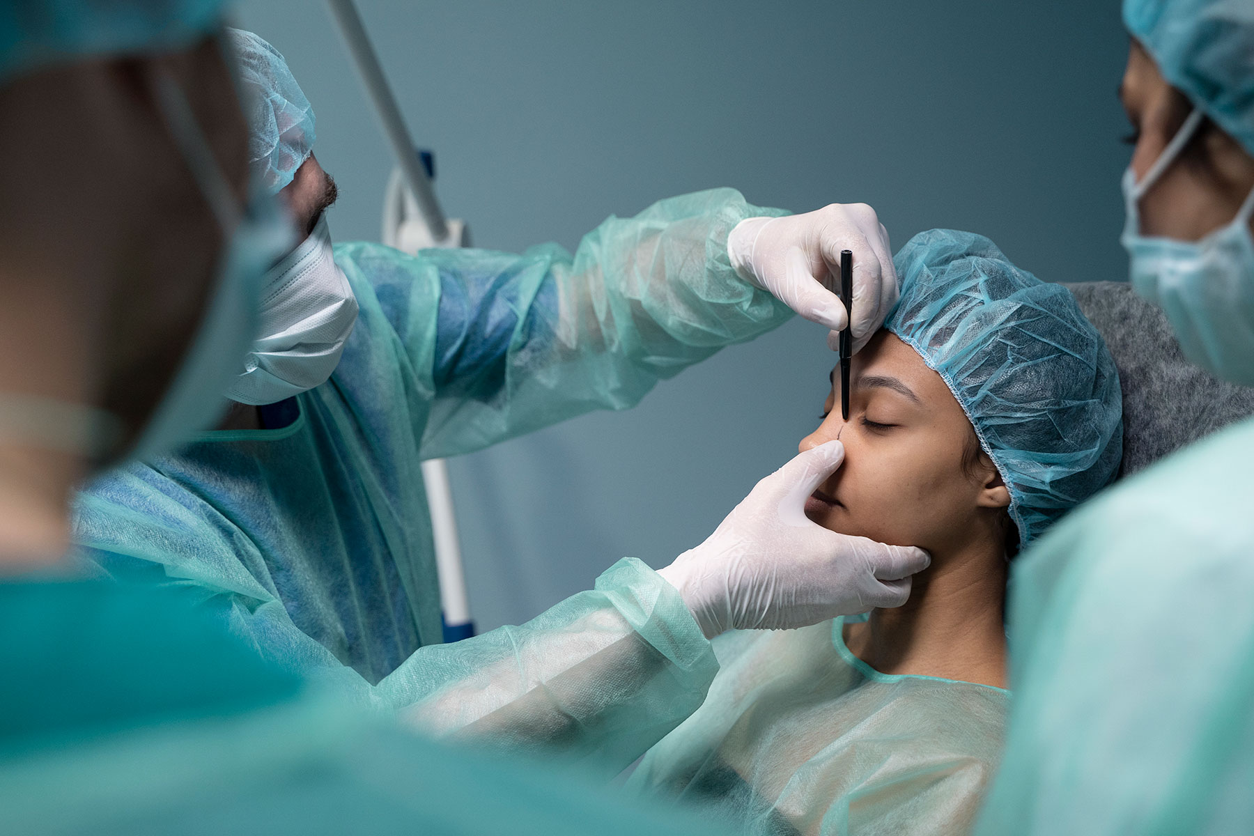 A medical team preparing for a nose surgery, marking the patient's nose with a pen while the patient is lying down, wearing a surgical gown and cap.