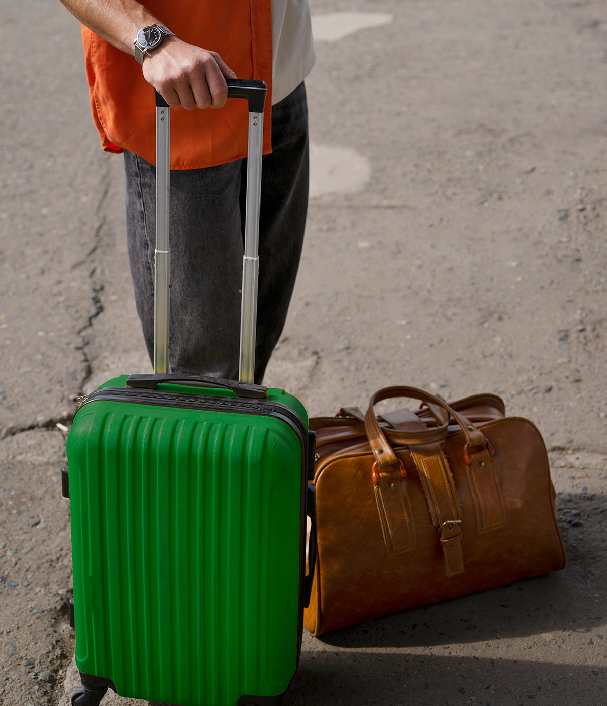 Person holding a green suitcase with one hand, next to a brown leather duffel bag, standing on a paved surface.