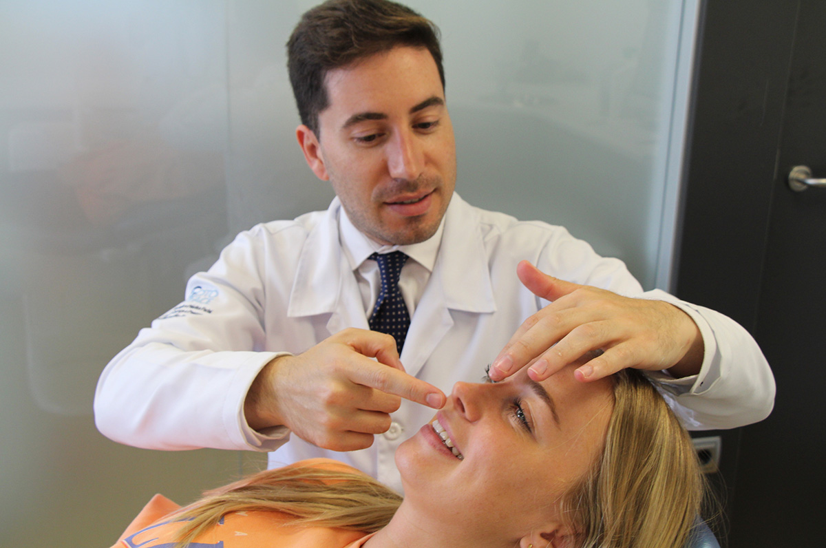 Doctor examining a patient's nose during a consultation, gently pointing at the area while the patient smiles, indicating a friendly and professional interaction.